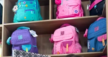 Prateleira de madeira com mochilas verde-água, roxas, rosa e azuis